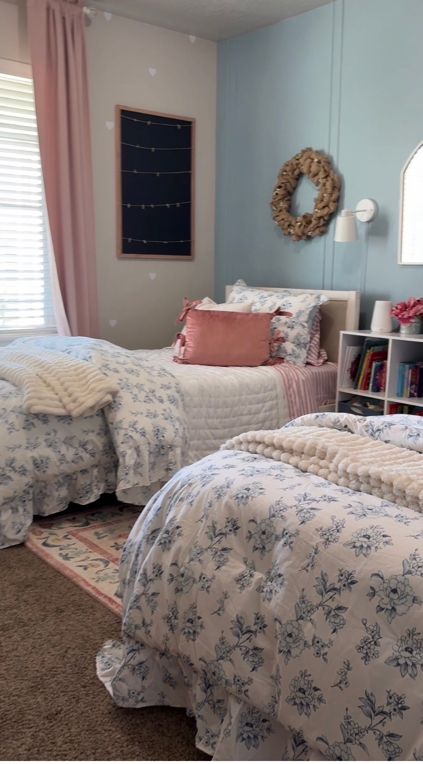 A cozy shared girls' bedroom with floral bedding, pink accents, blue board-and-batten accent wall, and organized bookshelf
